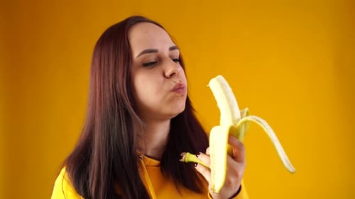 Close Up of Young Woman Eats Banana on Yellow Background
