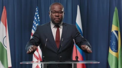 Man in Suit Speaking at Podium with Flags