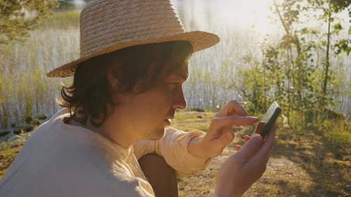 Young Man Surfs Internet on Smartphone Sitting on River Bank