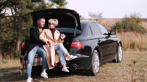 Stylish Couple Relaxing on Car in Grassy Field