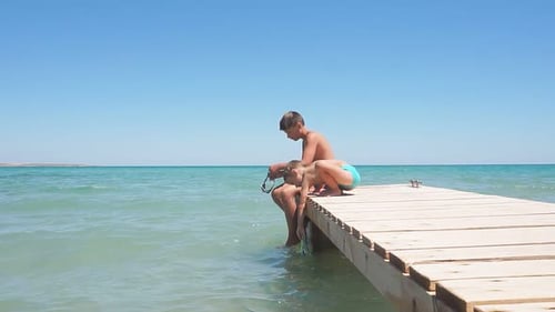 Close-up of Children Sitting on a Wooden Sea Pier, a Small Boy Touches the Water To Swim. Vacation