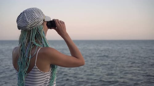 A Young Woman in a Marine Striped Dress Looks Through Binoculars at the Sea or Ocean
