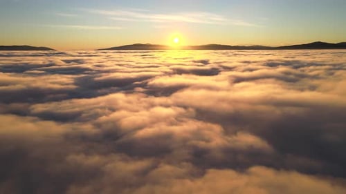 Luftaufnahme eines leuchtend gelben Sonnenaufgangs über weißen, dichten Wolken und fernen Bergen am Horizont.