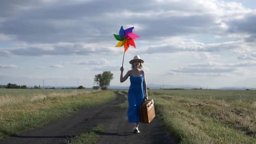 Girl in blue dress with suitcase and pinwheel on country road in summer.