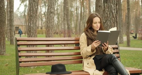 Young cute girl sitting in park on a bench and reading a book with black hat placed near her.