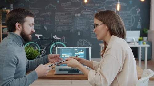 Businesswoman Talking To Colleague Discussing Information in Charts and Graphs on Laptop Screen and