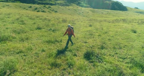 Flight Over Backpack Hiking Tourist Walking Across Green Mountain Field. Huge Rural Valley at Summer
