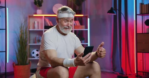 Hombre mayor sentado en el fitball en una habitación contemporánea por la noche y disfrutando de una videollamada