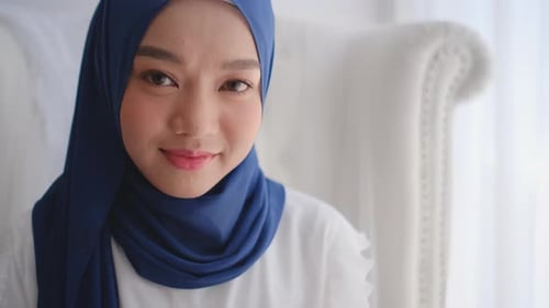 Muslimisches Mädchen schaut in die Kamera und lächelt, sitzt auch auf einem weißen Sofa in der Nähe von Glasfenstern