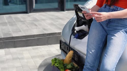 Nahaufnahme eines Mädchens benutzt Telefon beim Laden eines Elektroautos in der Nähe des Einkaufszentrums