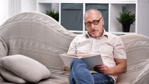 Focused Aged 70s Grandfather Reading Book Sitting on Couch
