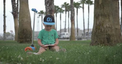 Young Boy Watches a Video on His Mobile Phone Sitting on the Grass in Park. Children and Modern