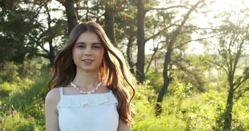 A Cheerful Teenage Girl in Light Dress Walks in a Clearing Between Green Trees in Summer Forest