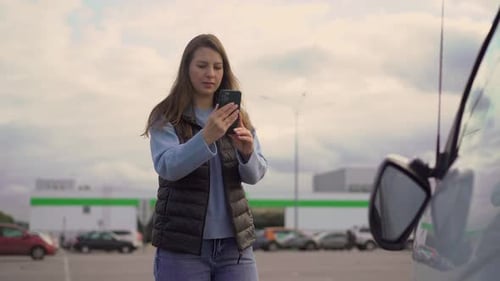 Woman Uses Smartphone Next to Car in Parking Lot