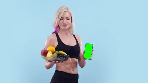 Beautiful Woman with an Athletic Body Holds Plate of Fruit and Phone with an App Green Screen