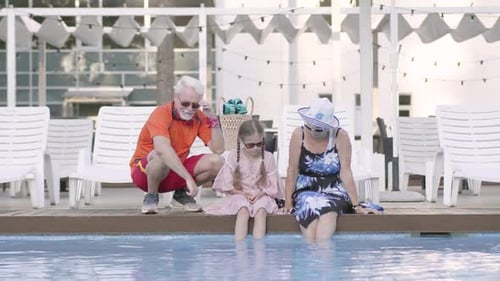 Happy Mature Couple and Their Little Granddaughter Sitting on the Edge of the Pool