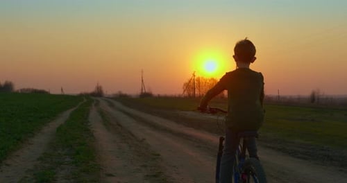 A Boy Is Riding a Bicycle on a Pathway at Sunset. Filming From the Back