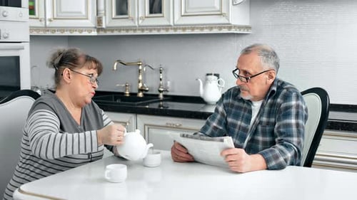 Mature Female Pouring Tea From Teapot Talking with Husband at Comfortable Kitchen