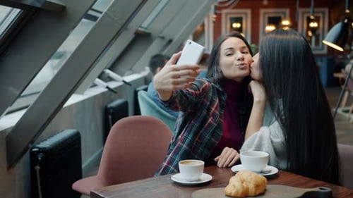 Charming Young Ladies Taking Selfie with Smartphone Camera Posing Kissing Having Fun in Cafe