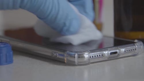 Close-up of Hand in Protective Blue Gloves Rubbing Smartphone Screen with Sanitizer. Unrecognizable
