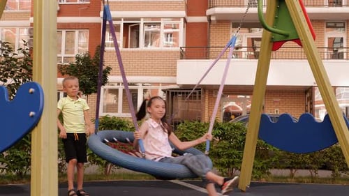 A Boy Rides a Girl on a Round Swing with a Net in the Yard on a Playground
