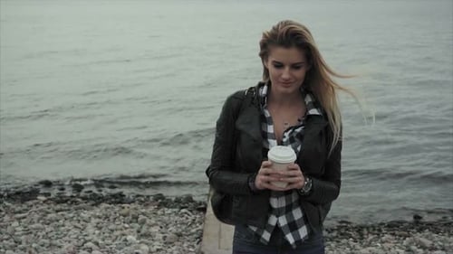 Pretty Young Blond Woman Holding a Cup of Coffee in Her Hands Near the Sea