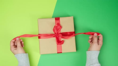 female hands untie red silk ribbon on gift box, green background