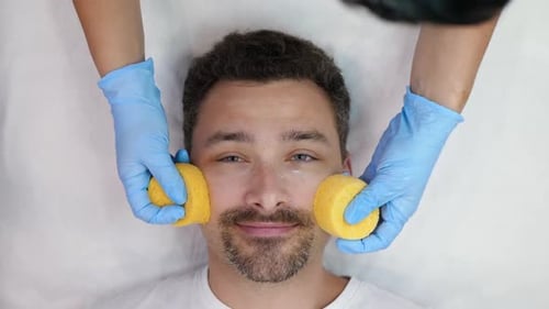 Man Receives Facial Cleansing Treatment with Yellow Sponges