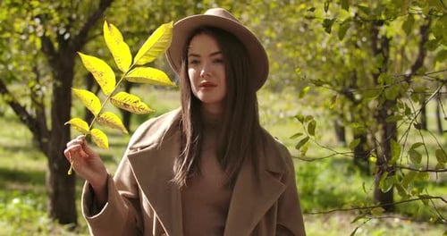 Thoughtful Caucasian Girl in Brown Hat Standing in the Autumn Park and Holding a Tree Branch