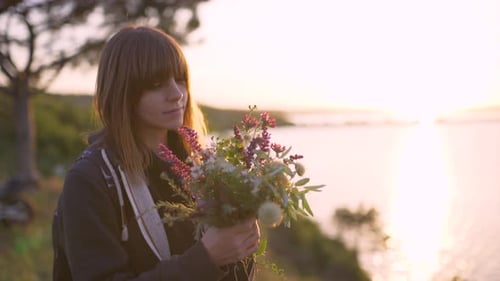 Beautiful Young Woman with a Bouquet of Wildflowers on the Coast at Sunset