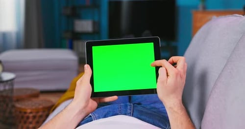 Young Man at Home Resting on a Couch Using with Green Mockup Screen Tablet Computer