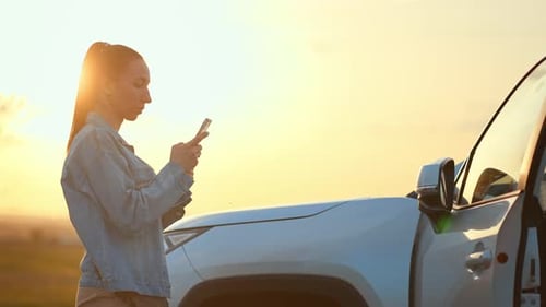Woman Using Phone Next to Car at Sunset