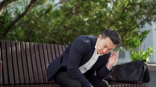 exhausted young asian business man office worker sitting on bench in city park outdoors.