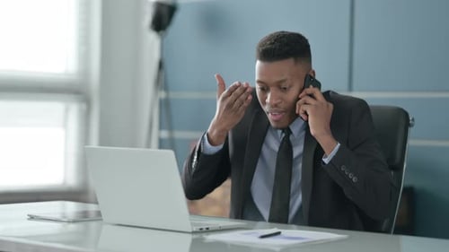 Angry African Businessman Talking on Smartphone while using Laptop in Office