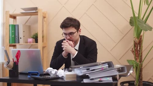 Office Worker Sits at Desk with Tired Look Palms Together and Looks Alternately at Laptop or Stack