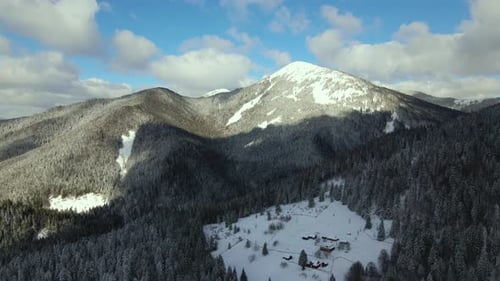 Aerial winter landscape with small rural houses between snow covered forest in cold mountains.