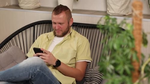Man Chatting Using Smartphone Sitting on Armchair at Home Types Message with Happy Smile