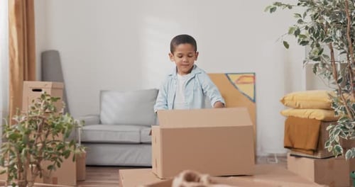 A Small Smiling Sweet Boy After Moving Into a New Apartment Looks Among the Boxes for His Toys Finds
