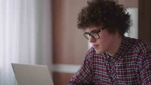 A Curly Man with a Serious Look Works at a Laptop Sitting in a Modern Kitchen