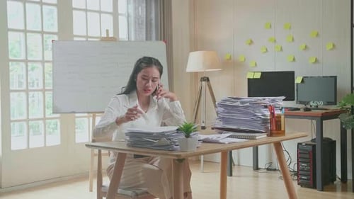 Asian Woman Talking On Phone And Taking Note While Working Hard With Documents At Office