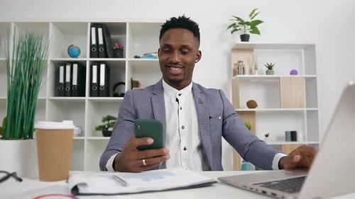 African american office manager sitting at his workplace and using smartphone