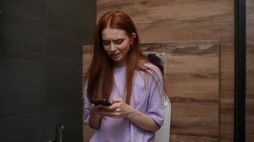 Cheerful Redhaired Young Woman Enjoying Using Mobile Phone Sitting on Toilet at Home Bathroom with