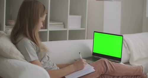 Schulmädchen sieht Online-Lektion auf Laptop mit Greenscreen für Chroma-Key-Technologie an und schreibt