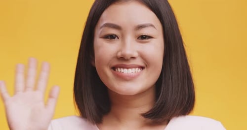 Nahaufnahme einer freundlichen asiatischen Dame, die mit einer Hallo-Geste mit der Hand winkt und lächelt, orangefarbener Studio-Hintergrund
