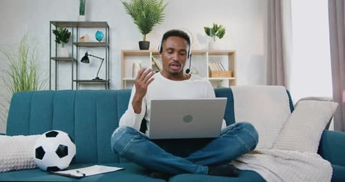 Hombre con auriculares sentado en el sofá de casa y celebrando una videoconferencia en una computadora portátil