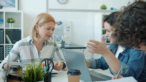 Cheerful Women Conversing and Using Laptop Working Indoors in Light Office