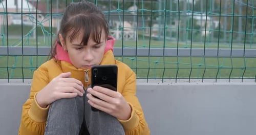 Child Using Phone Sitting Near Green Mesh Fence
