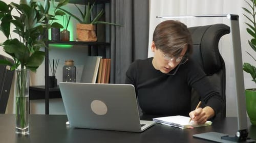 Multitasking female is talking on phone, taking notes, typing on keyboard sitting in modern office