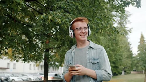 Happy Man in Headphones Walking in Park Listening To Music Using Smartphone