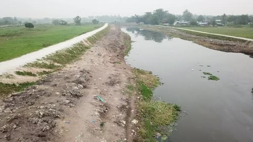 Aerial fly toward the dark polluted river with rubbish around the river bank